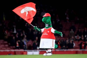 O mascote do Arsenal, Gunnersaurus, dentro de campo com a bandeira do clube na mão direita.